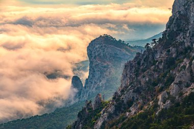 Kayalık dağların ve vadinin üzerinde bulutların ve sislerin içinde görkemli turuncu günbatımı. Kremalı sis dağ vadisini gün batımıyla kapladı. Kırım Dağları üzerinde puslu gün batımı. Ai-Petri, Kırım.
