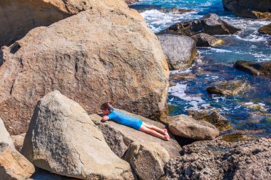 Mayo giymiş küçük bir çocuk açık güneşli bir günde deniz kenarındaki kayalıklarda dalgalarla şeffaf bir denizin arka planında güneşleniyor..