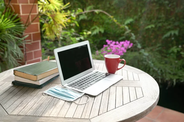 Laptop, face mask, books and red cup on top of table next to garden. Study online during pandemic concept.