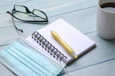 Sarı kalemli, maskeli, mavi ahşap masadaki gözlüklü ve kahveli defteri aç..