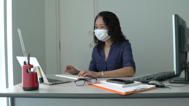顔のマスクを身に着けているビジネスマンの女性 彼女の作業スペースでマルチタスク コンピュータを入力し 彼女の携帯電話でチェック — ストック動画