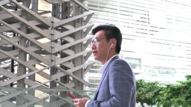 Asiatisk Affärsman Hörlurar Håller Sin Mobiltelefon Och Pratar Offentlig Miljö — Stockvideo