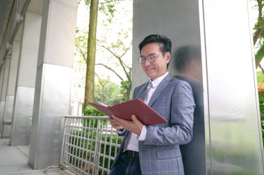 Asyalı işadamı inşaat direğine yaslanıyor, günlüğüne bakarken gülümsüyor..