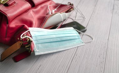 Maskeli kırmızı kadın çantası, el dezenfektanı, kulaklık ve ahşap masada akıllı telefon..
