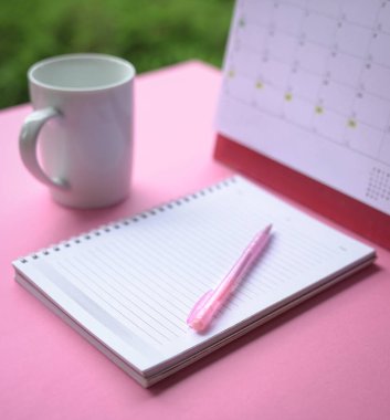 Defterin üstüne pembe kalem, takvim ve arka planda bir fincan kahve. Seçici odak.