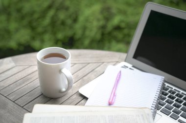 Bir fincan kahve, not defteri, açık kitap, masa başında kalem ve bilgisayar, arka planda yeşil bitkiler..