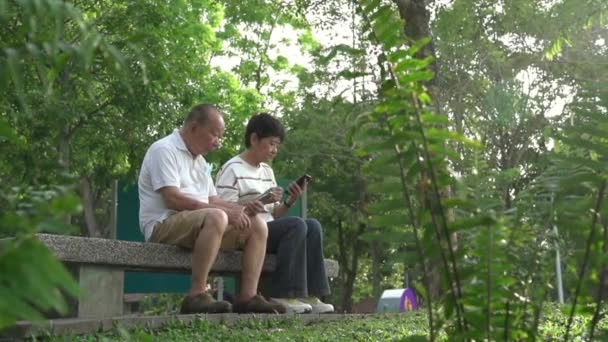 Casal Asiático Sênior Sentado Banco Parque Lendo Surfando Seus Telefones — Vídeo de Stock