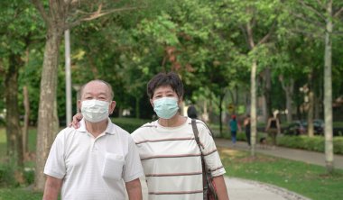 Yeşil bir parkta yürüyen, yüz maskesi takan yaşlı Asyalı çift..