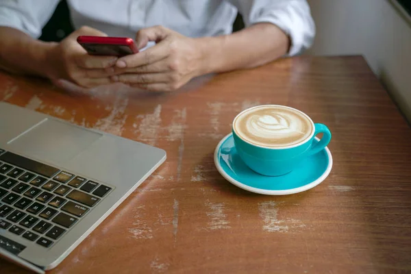 Man reading phone with computer and cup of coffee on table.  Close up view on cup of coffee in blue cup.