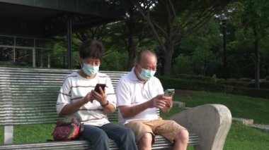 Yüz maskeli yaşlı bir Asyalı çift bankta oturuyor ve telefonlarını mesajlaşmak, ailesiyle iletişim kurmak ve haberleri yakalamak için kullanıyorlar. Açık hava parkı ayarları.