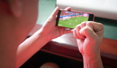 Spor fanatiği, cep telefonundan spor haberlerini izlerken yumruğunu havaya kaldırıyor. Görüntüyü kapat.