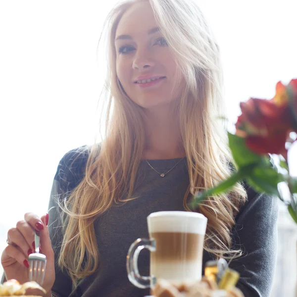 Chica, comiendo un pedazo de pastel y una taza de café con leche en un café en el fondo borroso de la ciudad — Foto de Stock