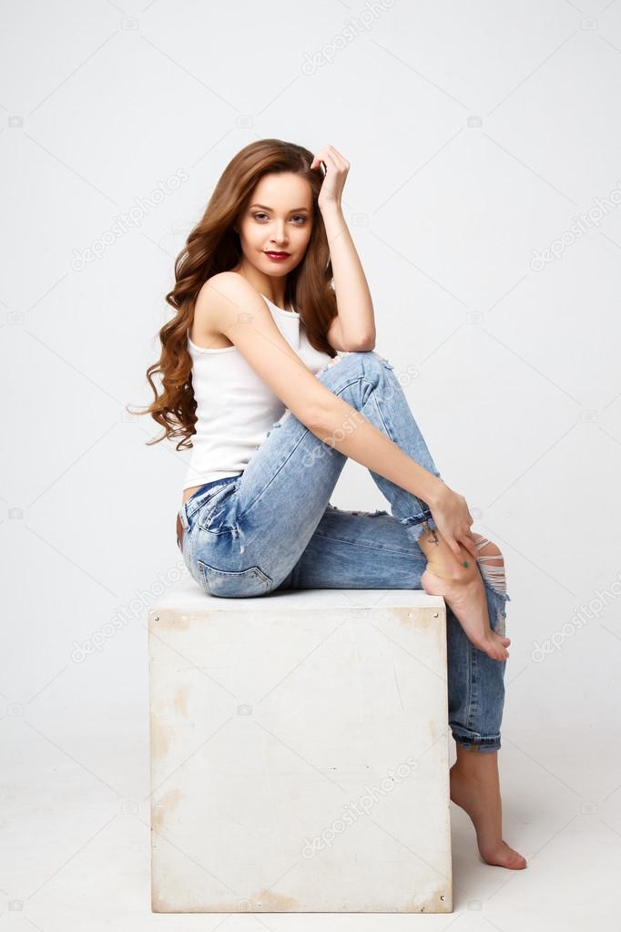Close-up portrait of beautiful young woman with gorgeous hair and natural makeup sitting on a white cube. Fashion beauty photo, casual jeans style