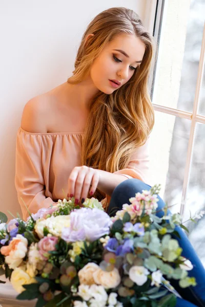 Mujer rubia joven con un ramo de flores sentado cerca de la ventana. Retrato tierno en una mañana — Foto de Stock