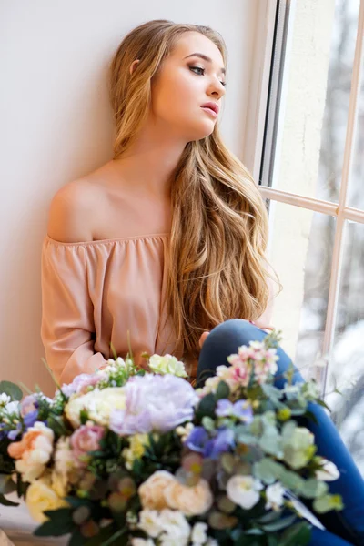 Pencerenin yanında elinde bir buket çiçekle oturan genç sarışın kadın. Bir sabah şefkatli bir portre