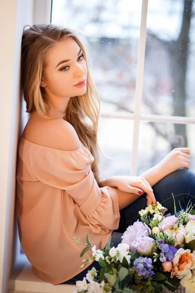 Mujer rubia joven con un ramo de flores sentado cerca de la ventana. Retrato tierno en una mañana — Foto de Stock