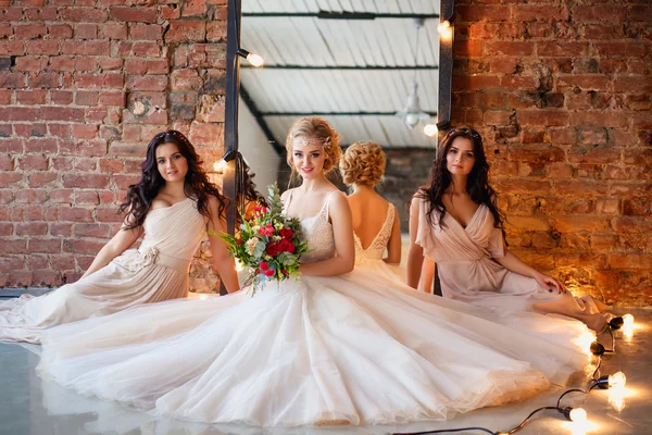 Hermosa novia rubia en vestido de novia de lujo y bonitas damas de honor gemelas en vestidos similares en una mañana en un loft espacio con un espejo y guirnalda de lámparas. Moda boda moderna foto . —  Fotos de Stock