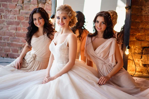 Hermosa novia rubia en vestido de novia de lujo y bonitas damas de honor gemelas en vestidos similares en una mañana en un loft espacio con un espejo y guirnalda de lámparas. Moda boda moderna foto . — Foto de Stock