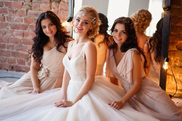 Hermosa novia rubia en vestido de novia de lujo y bonitas damas de honor gemelas en vestidos similares en una mañana en un loft espacio con un espejo y guirnalda de lámparas. Moda boda moderna foto . —  Fotos de Stock