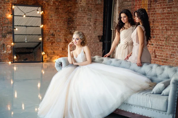Mooie blonde bruid in luxe trouwjurk en mooie tweeling bruidsmeisjes in soortgelijke jurken in een ochtend in een zolderruimte met een spiegel en Garland van lampen. Mode moderne bruiloft foto. — Stockfoto