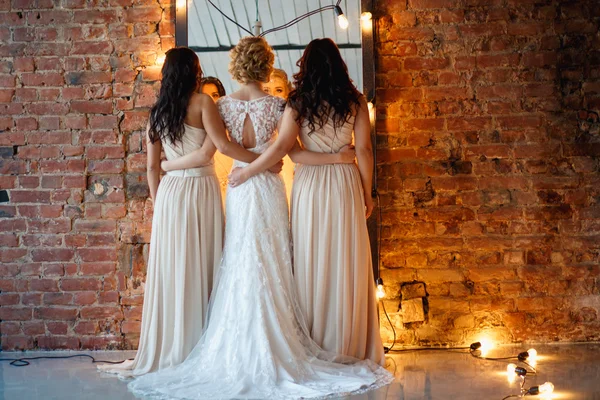 Belle mariée blonde en robe de mariée de luxe et jolies demoiselles d'honneur jumelles dans des robes similaires le matin dans un espace loft avec un miroir et une guirlande de lampes. Mode moderne photo de mariage . — Photo