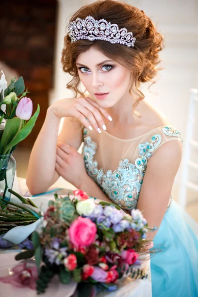 Hermosa joven en un vestido azul de lujo sentado junto a una mesa con un ramo de flores —  Fotos de Stock