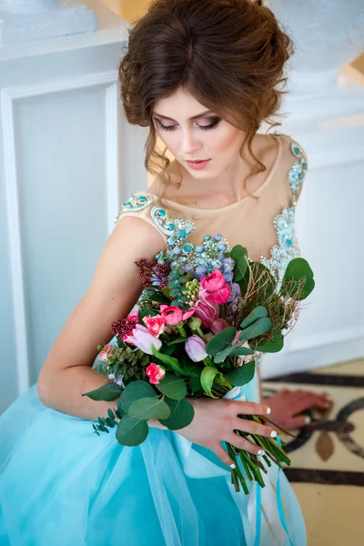 Hermosa joven en un vestido azul de lujo en un elegante interior con un ramo de flores — Foto de Stock