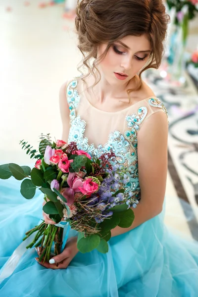 Hermosa joven en un vestido azul de lujo en un elegante interior con un ramo de flores — Foto de Stock