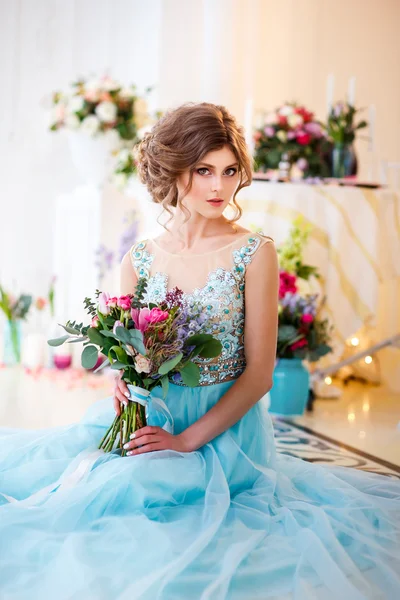 Hermosa joven en un vestido azul de lujo en un elegante interior con un ramo de flores — Foto de Stock