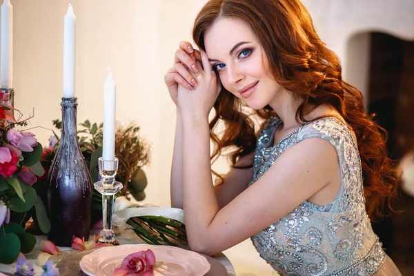 Menina bonita em um vestido azul de luxo sentado ao lado de uma mesa com um buquê de flores — Fotografia de Stock