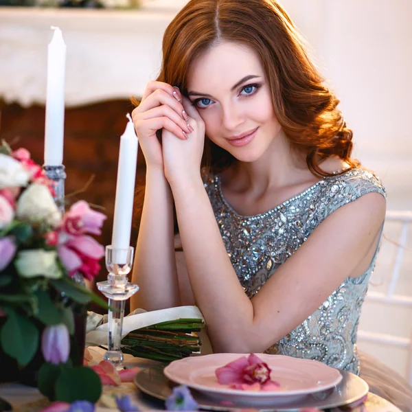 Schöne junge Dame in einem luxuriösen blauen Kleid sitzt an einem Tisch mit einem Blumenstrauß — Stockfoto