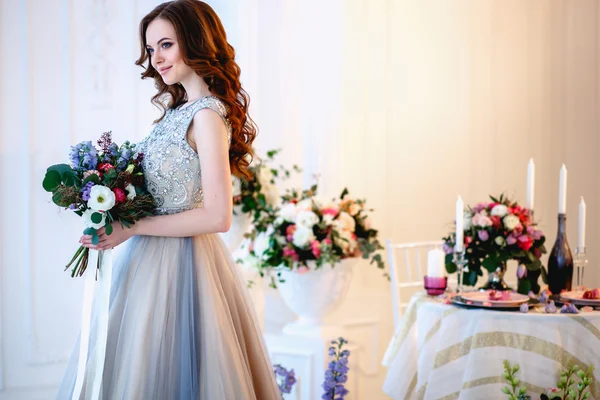 Hermosa joven en un vestido de lujo en un elegante interior con un ramo de flores — Foto de Stock