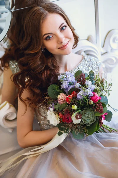 거울 근처에 앉아 꽃다발과 우아한 인테리어의 고급 드레스를 입은 아름다운 젊은 아가씨 — 스톡 사진