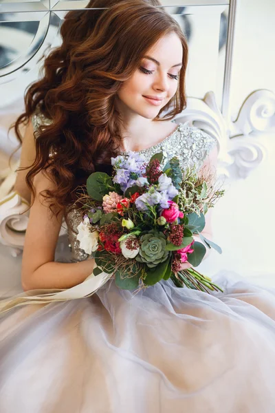 Hermosa joven en un vestido de lujo en un elegante interior con un ramo de flores sentado cerca del espejo — Foto de Stock