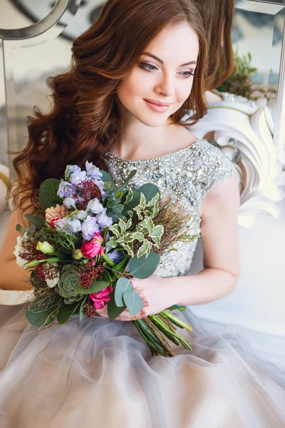 Hermosa joven en un vestido de lujo en un elegante interior con un ramo de flores sentado cerca del espejo — Foto de Stock