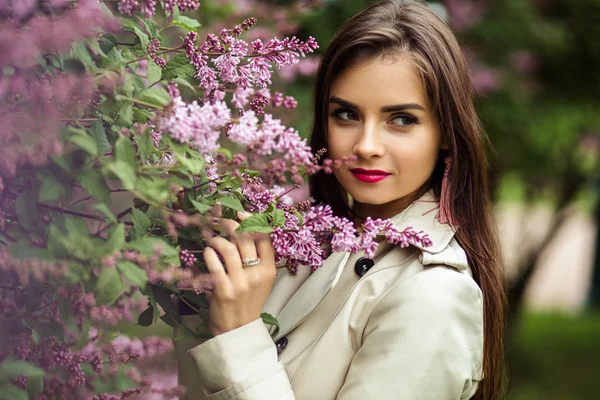 Bela jovem mulher em um casaco de trincheira perto de floração lilás — Fotografia de Stock