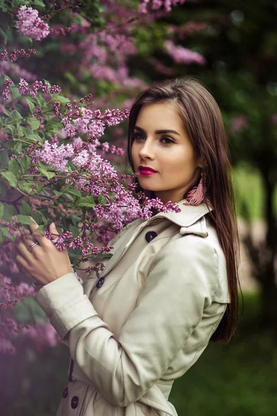 Bela jovem mulher em um casaco de trincheira perto de floração lilás — Fotografia de Stock