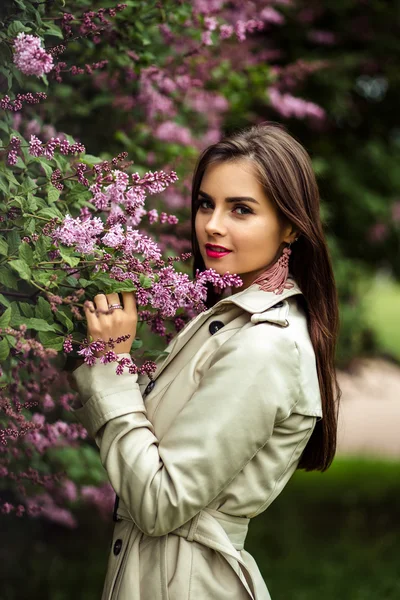 Bela jovem mulher em um casaco de trincheira perto de floração lilás — Fotografia de Stock