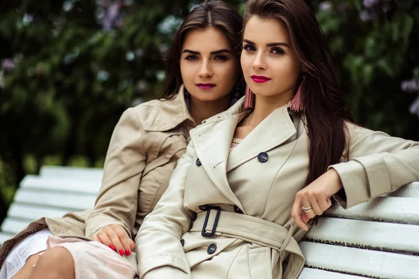 Dos hermosas gemelas mujeres jóvenes en abrigos de trinchera cerca de la floración lila —  Fotos de Stock