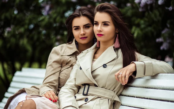 Two beautiful twins young women in trench coats near blooming lilac — Stock Photo, Image