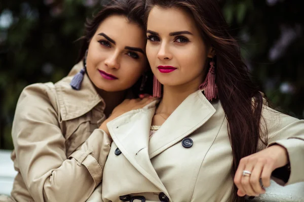 Deux belles jumelles jeunes femmes en trench manteaux près de fleurs lilas — Photo