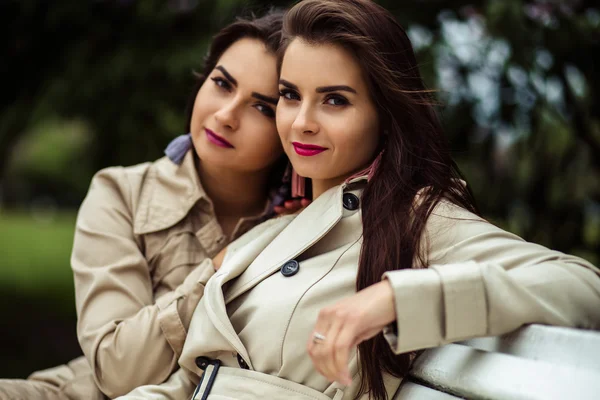 Dos hermosas gemelas mujeres jóvenes en abrigos de trinchera cerca de la floración lila — Foto de Stock