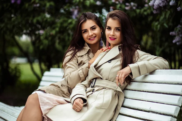 Deux belles jumelles jeunes femmes en trench manteaux près de fleurs lilas — Photo