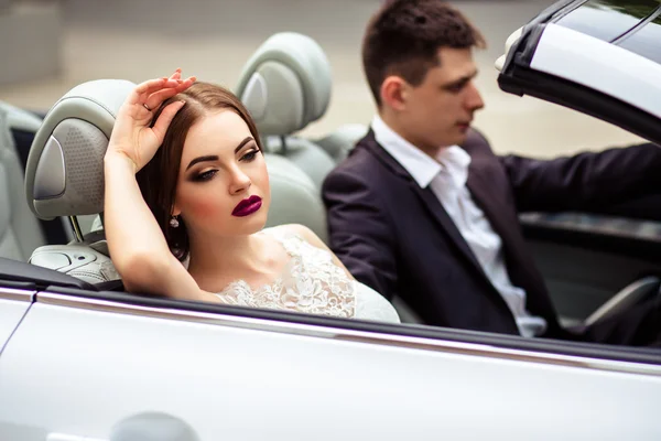 Gorgeous bride with fashion makeup and hairstyle in a luxury wedding dress with handsome groom near white cabriolet car — Stock Photo, Image