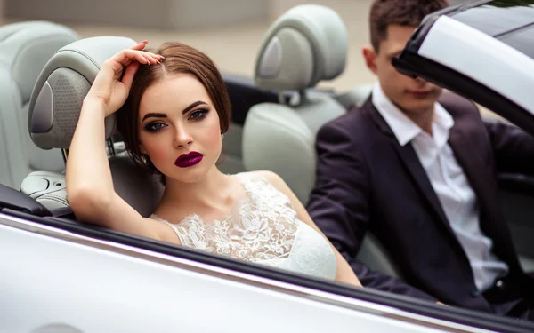 Gorgeous bride with fashion makeup and hairstyle in a luxury wedding dress with handsome groom near white cabriolet car — Stock Photo, Image