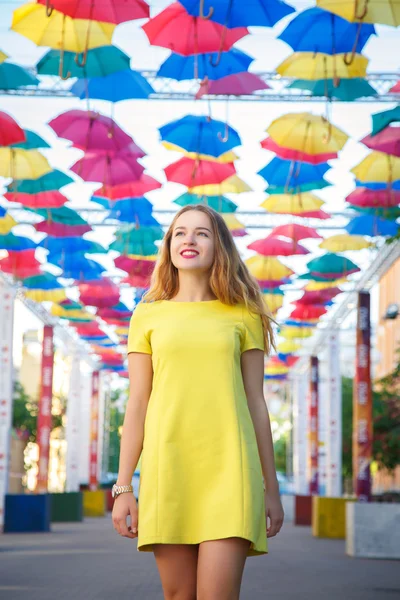 Krásná dívka ve žlutých šatech pózuje na pozadí barevné deštníky alej — Stock fotografie