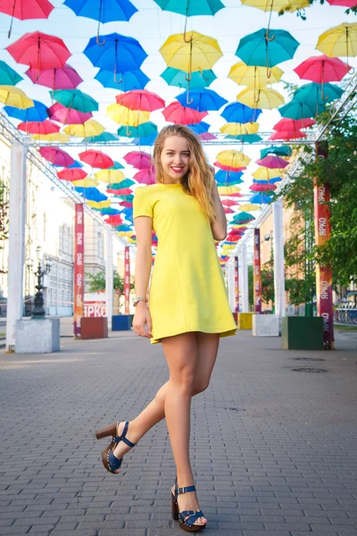Krásná dívka ve žlutých šatech pózuje na pozadí barevné deštníky alej — Stock fotografie