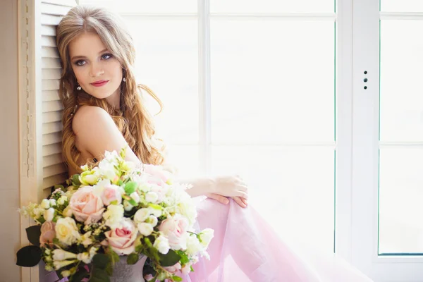 Retrato de niña bonita en un vestido dulce y pelo largo y rubio sentado en una ventana con composición de flores —  Fotos de Stock