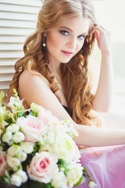 Retrato de menina bonita em um vestido doce e longo cabelo loiro sentado em uma janela com composição de flores — Fotografia de Stock