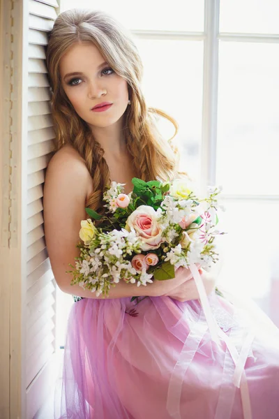 Портрет красивої молодої дівчини в солодкій сукні і довге світле волосся, що сидить на вікні з квітковою композицією — стокове фото